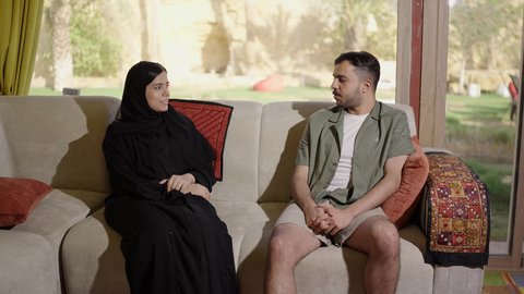قضاء أوقات ممتعة، زوجان عربيان خليجيان سعوديان يستمتعان بتبادل الحديث على الأريكة في غرفة المعيشة، نقاشات وحوارات، أجواء عائلية سعيدة، ايماءات السعاده و السرور