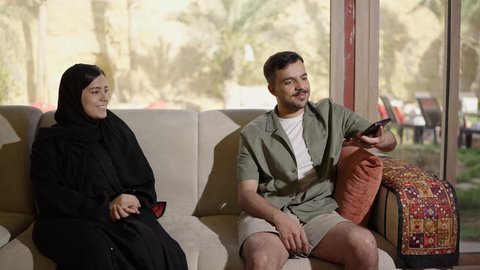 مفهوم المودة والالفة ، قضاء أوقات ممتعة، زوجان عربيان خليجيان سعوديان مبتسمان يجلسان في غرفة المعيشة ، رجل عربي يرتدي الزي الكاجوال يقوم بإلتقاط صورة سيلفي برفقة زوجته عبر الهاتف المحمول بايماءات السعادة و السرور