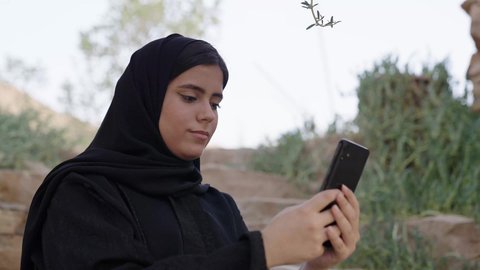 قضاء وقت ممتع فى الخارج, تصوير مقرب لامراة عربيه خليجيه سعودية ترتدي الحجاب و العباءة السوداء تجلس فى الهواء الطلق تستخدم الهاتف المحمول ، استخدام البرامج والتطبيقات الالكترونية، التسوق الالكتروني عبر الانترنت، تصفح المواقع الالكترونية والتواصل الاجتماعي