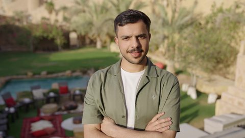 الأناقة والاعتناء بالمظهر الخارجي، مفهوم الرجولة والوسامة، الوقوف بأيدي متقاطعة بإيماءات الثقة بالنفس، تصوير مقرب لشاب عربي خليجي سعودي يرتدي الزي الكاجوال يقف في الهواء الطلق و ينظر الى الكاميرا بتعابير السرور والبشاشة