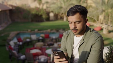 الاستمتاع بقضاء وقت ممتع فى الخارج، تصفح المواقع الالكترونية والتواصل الاجتماعي, تصوير لرجل عربي خليجي سعودي يرتدي الزي الكاجوال يجلس فى الهواء الطلق يستخدم الهاتف المحمول