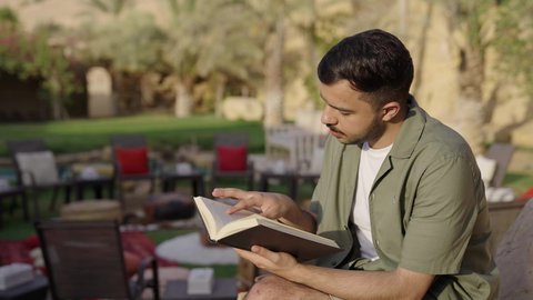Spending free time on education and learning, practicing the hobby of reading, the concept of culture, awareness, and self-learning, a depiction of a young Arab Gulf Saudi man dressed in casual attire reading and studying outdoors.