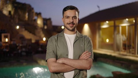 الوقوف بأيدي متقاطعة بإيماءات الثقة بالنفس، شاب عربي خليجي سعودي يرتدي الزي الكاجوال يقف في الهواء الطلق و ينظر الى الكاميرا بتعابير السرور والبشاشة، الاهتمام بالمظهر الخارجي والأناقة، مفهوم الرجولة والوسامة