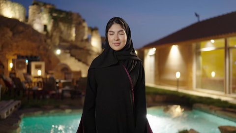 ملابس وأزياء محجبات عصرية راقية، النظر الى الكاميرا بتعابير السرور، الملامح العربية الأصيلة، مفهوم الثقة بالنفس والاهتمام بالمظهر الخارجي، تصوير لامرأة عربية خليجية سعودية مبتسمة ترتدي الحجاب والعباءة تقف بأيدي متقاطعة فى الهواء الطلق