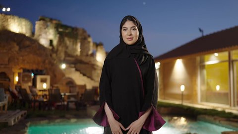 النظر الى الكاميرا بتعابير السرور، الملامح العربية الأصيلة، ملابس وأزياء محجبات عصرية راقية، مفهوم الثقة بالنفس والاهتمام بالمظهر الخارجي، تصوير لامرأة عربية خليجية سعودية مبتسمة ترتدي الحجاب والعباءة تقف بأيدي متقاطعة فى الهواء الطلق