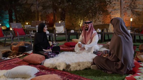 ممارسة ألعاب ممتعة لقضاء وقت الفراغ، المتعة والسعادة في أحد الشاليهات في العطلة الصيفية، تصوير  لشاب عربي خليجي سعودي يرتدي الثوب الابيض و الشماغ يجلس فى الهواء الطلق برفقة صديقاته و يقوم بشرح لعبة الورق