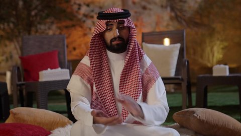 Spending enjoyable time engaging in fun activities and games, joy and happiness in one of the chalets during the summer vacation, a close-up shot of a young Arab Gulf Saudi man wearing a white thobe and a ghutrah sitting outdoors and explaining a card game.