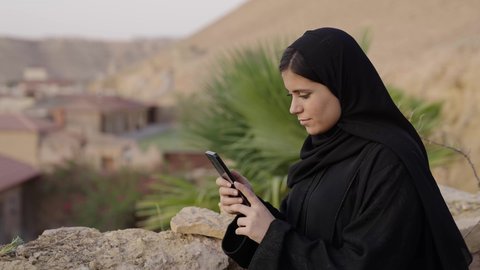 Using electronic programs and applications, browsing websites and social media, spending enjoyable time outdoors, photographing an Arab Gulf Saudi woman wearing a hijab and a black abaya sitting outdoors using a mobile phone, online shopping.