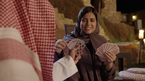 التسلية والترفيه والاستمتاع باللعب الجماعي، خلط الأوراق بحركات احترافية، تصويرلامراه عربيه خليجيه سعودية ترتدي العباءه و الحجاب تقوم بالعب لعبه الورق برفقه زوجها ، ايماءات السعاده و السرور