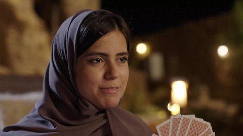 تسليه وترفيه مع العائله،  ايماءات السعادة و السرور، الإحتراف في خلط الورق, مفهوم التركيز و التحدي, التخطيط والتفكير, قضاء وقت ممتع ، تصوير مقرب لامراة عربيه خليجيه سعودية ترتدي الحجاب و العباءة تجلس فى الهواء الطلق و تقوم بالعب لعبه الورق