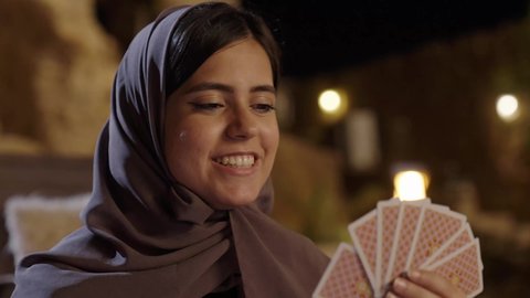 أنشطه ترفيهية فى الخارج،  ايماءات السعادة و السرور، الإحتراف في خلط الورق, مفهوم التركيز و التحدي, التخطيط والتفكير, قضاء وقت ممتع, تسليه وترفيه مع العائله ، تصوير مقرب لامراة عربيه خليجيه سعودية ترتدي الحجاب و العباءة تجلس فى الهواء الطلق و تقوم بالعب لعبه الورق