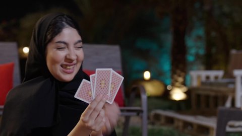 ايماءات السعادة و السرور، الإحتراف في خلط الورق, مفهوم التركيز و التحدي, التخطيط والتفكير, أنشطه ترفيهية فى الخارج, قضاء وقت ممتع, تسليه وترفيه مع العائله ، تصوير مقرب لامراة عربيه خليجيه سعودية ترتدي الحجاب و العباءة تجلس فى الهواء الطلق و تقوم بالعب لعبه الورق