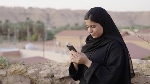 تصفح المواقع الالكترونية والتواصل الاجتماعي، قضاء وقت ممتع فى الخارج, تصوير لامراة عربيه خليجيه سعودية ترتدي الحجاب و العباءة السوداء تجلس فى الهواء الطلق تستخدم الهاتف المحمول ، استخدام البرامج والتطبيقات الالكترونية، التسوق الالكتروني عبر الانترنت، عرض شاشة خضراء عبر الجوال