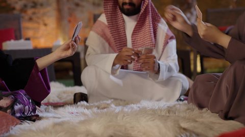 مفهوم الترفيه العائلي، تصوير مقرب لعائله عربيه خليجيه سعودية يرتدون الزي التقليدي يجلسون على الأرض يتبادلون اللعب بأوراق الكوتشينة، قضاء وقت ممتع مع الأسرة والأصدقاء