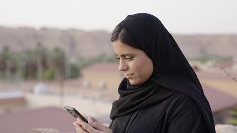 عرض شاشة خضراء عبر الجوال، قضاء وقت ممتع فى الخارج, تصوير مقرب من الجانب لامراة عربيه خليجيه سعودية ترتدي الحجاب و العباءة السوداء تجلس فى الهواء الطلق تستخدم الهاتف المحمول ، استخدام البرامج والتطبيقات الالكترونية، التسوق الالكتروني عبر الانترنت، تصفح المواقع الالكترونية والتواصل الاجتماعي