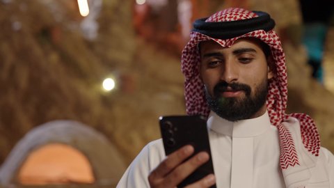 استخدام جهاز تقني حديث متطور، تصوير مقرب لرجل عربي خليجي سعودي يرتدي الثوب الابيض و الشماغ يجلس فى الهواء الطلق يمسك بيده الهاتف المحمول، تصفح وسائل التواصل الاجتماعي، قضاء وقت ممتع فى الخارج