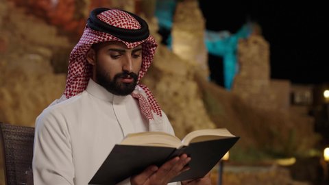 مفهوم التثقيف والبحث العلمي، هواية المطالعة والاستكشاف، قضاء وقت الفراغ بالتعلم والثقافة، رجل عربي خليجي سعودي يرتدي الثوب الابيض و الشماغ يجلس فى الهواء الطلق و يقوم بقراءة كتاب