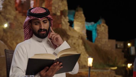قضاء أوقات ممتعة في قراءة الكتب المفيدة للتعلم والتثقف، تبادل الأحاديث و الأخبار الجيدة، إجراء مكالمة هاتفية صوتية عبر الجوال، تصوير لرجل عربي سعودي خليجي يرتدي الثوب الابيض و الشماغ  يجلس في الهواء الطلق يمسك بيديه كتاب ويتحدث عبر الهاتف المحمول، إيماءات السعادة بالتواصل مع الأصدقاء والأقارب