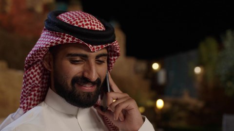 تبادل الأحاديث و الأخبار الجيدة، إجراء مكالمة هاتفية صوتية عبر الجوال، تصوير مقرب رجل عربي سعودي خليجي يرتدي الثوب الابيض و الشماغ  يجلس في الهواء الطلق  ويتحدث عبر الهاتف المحمول، إيماءات السعادة بالتواصل مع الأصدقاء والأقارب