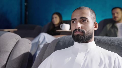 ايماءات السعاده و السرور، دور العرض الحديثة في المملكة العربية السعودية، القيام بنشاطات ترفيهية ممتعة, تصوير مقرب لرجل عربي خليجي سعودي يرتدي الثوب الابيض يجلس  في قاعة السينما يتابع الفيلم بتركيز, مشاهدة الأفلام والمسرحيات والعروض المتنوعة