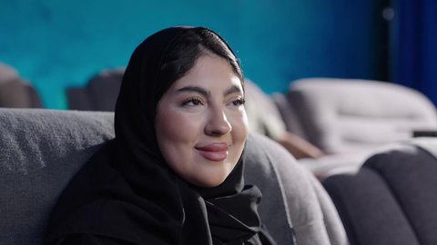 Modern cinemas in the Kingdom of Saudi Arabia, engaging in enjoyable recreational activities, a close-up shot of a Saudi Gulf Arab woman wearing a hijab sitting in a cinema hall and watching the movie attentively, watching films, plays, and various shows, gestures of happiness and joy.