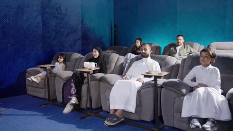 Watching movies and theatrical performances in the cinema, a Saudi Arabian Gulf Arab family sitting in the cinema hall, expressions indicating happiness and engagement, spending enjoyable family time, background of the cinema screening room, screening venues.