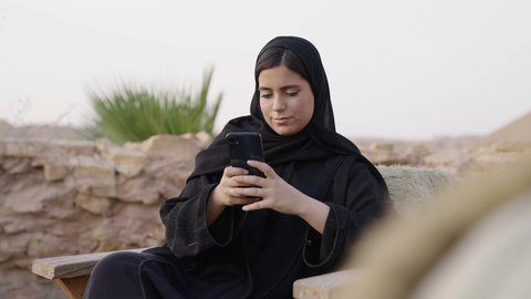 التسوق الالكتروني عبر الانترنت، تصفح المواقع الالكترونية والتواصل الاجتماعي، قضاء وقت ممتع فى الخارج, تصويرلامراة عربيه خليجيه سعودية ترتدي الحجاب و العباءة السوداء تجلس فى الهواء الطلق تستخدم الهاتف المحمول ، استخدام البرامج والتطبيقات الالكترونية