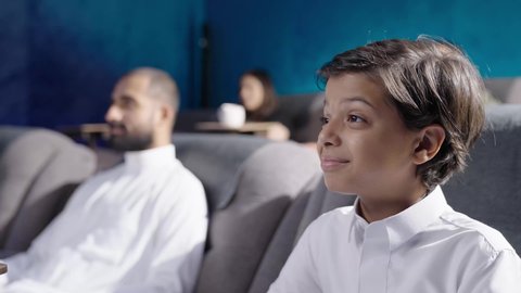 ايماءات الاندماج و التركيز، الاستمتاع بوقت الفراغ في مشاهدة الأفلام داخل السينما، تصوير مقرب من الجانب لولد عربي خليجي سعودي يرتدي الثوب الابيض يجلس داخل قاعه السينما، الاستمتاع بمشاهده فيلم