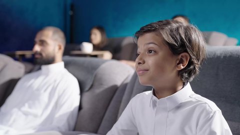 Enjoying free time watching movies in the cinema, a close-up shot of a Saudi Gulf Arab boy wearing a white thobe sitting inside the cinema hall, enjoying watching a movie, gestures of immersion and enjoyment.