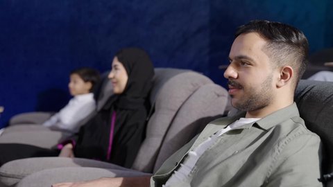 Weekend recreational activities, happy family atmosphere, a Saudi Gulf Arab family sitting on a chair watching a cinema screen, watching a movie, gestures of happiness and joy.