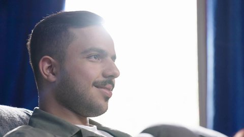 النظر الى شيء ما، مفهوم الرجولة والاهتمام بالمظهر الخارجي، تصوير من الجانب لرجل عربي خليجي سعودي يرتدي الزي الكاجوال يجلس بجوار نافذة مشرقة في غرفة مشمسة، ايماءات السعادة و السرور