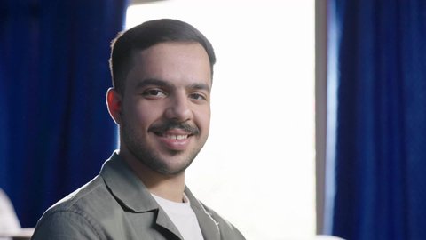 The concept of elegance and masculinity, an Arab Gulf Saudi man wearing casual attire sitting next to a bright window in a sunny room, looking at the camera with gestures of happiness and joy.