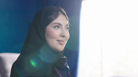 النظر الى شيء ما، مفهوم الأنوثة والنعومة، الاهتمام بالمظهر الخارجي والأناقة، تصويرمقرب من الجانب لامراة عربية خليجية سعودية تجلس بجوار نافذة مشرقة في غرفة مشمسة ترتدي الحجاب و العباءة السوداء تجلس بجانب نافذه في غرفه مشمسه