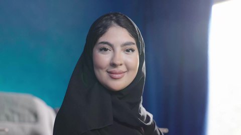 الاهتمام بالمظهر الخارجي والأناقة، مفهوم الأنوثة والنعومة، تصويرمقرب لامراة عربية خليجية سعودية تجلس بجوار نافذة مشرقة في غرفة مشمسة ترتدي الحجاب و العباءة السوداء، النظرالى الكاميرا بايماءات السعادة و السرور