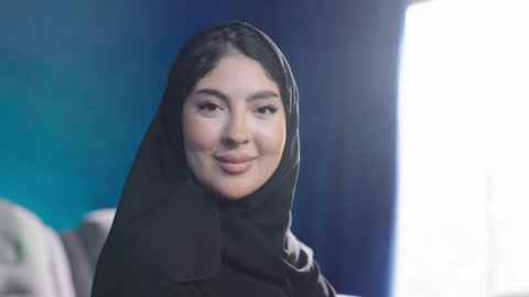 مفهوم الأنوثة والأناقة والشياكة والنعومة، تصويرمقرب لامراة عربية خليجية سعودية تجلس بجوار نافذة مشرقة في غرفة مشمسة ترتدي الحجاب و العباءة السوداء تجلس بجانب نافذه في غرفه مشمسه، النظرالى الكاميرا بايماءات السعادة و السرور