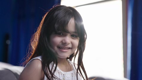 أجواء طفولة وبهجة داخلية، تصويرمقرب لبنت عربيه خليجيه سعودية مبتسمه ترتدي الزي الكاجوال تجلس بجانب نافذه في غرفه مشمسه، النظرالى الكاميرا بايماءات السعادة و السرور