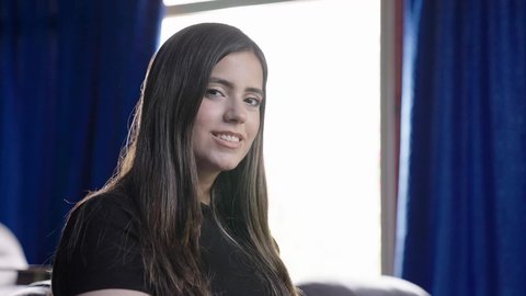 النظر الي الكاميرا بايماءات السعادة و السرور، اجواء هادئه و تامل عميق ، تصوير لفتاه عربيه خليجيه سعوديه ذات ملامح وجه وشعر بني ناعم ترتدي الزي الكاجوال تجلس بجانب نافذه في غرفه مشمسه