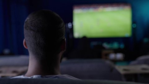 Challenge, encouragement, and strength, expressions of passion and enthusiasm for winning, harmony and interaction with the competition, raising hands up in expressions of victory, a shot from behind of a young Arab Saudi Gulf man wearing a white thobe watching matches and sports games, gestures of joy, delight, and happiness.