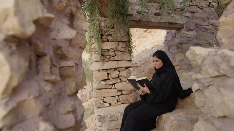 قراءة الروايات والقصص، ايماءات التركيز والاستمتاع، تمضية أوقات الفراغ بالتعلم والتثقيف الذات، ثقافة القراءة في أجواء تاريخية عربية, امرأة عربيه خليجيه سعودية ترتدي العباءة السوداء تستمتع بالقراءة بين الآثار القديمة