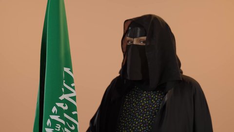 Showing loyalty to the country, our beginning day February 22, Saudi National Day September 23, wearing traditional dress on a national occasion, Flag Day March 11, close-up portrait of a Saudi Arabian Gulf woman wearing a burqa and abaya holding the Kingdom's flag and looking at the camera with proud gestures, floral background