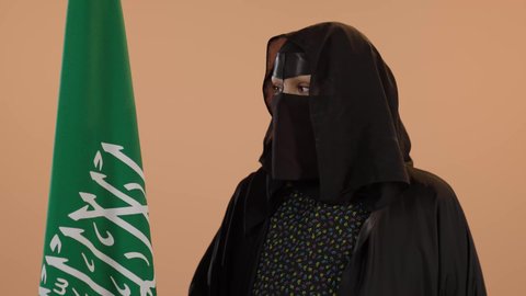 Wearing traditional costume on a national occasion, Flag Day March 11, showing loyalty to the country, Badina Day February 22, Saudi National Day September 23, close-up portrait of a Saudi Arabian Gulf woman wearing a burqa and abaya holding the Kingdom's flag and looking at the camera with proud gestures, floral background
