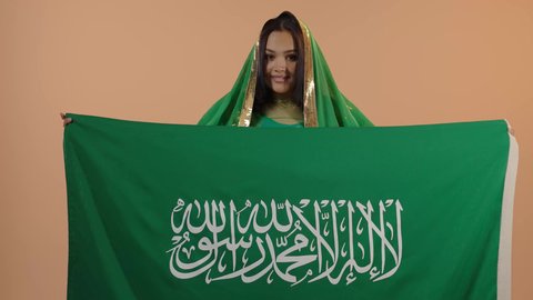 التباهي بإنجازات الوطن، راية التوحيد وسيف القوة، اليوم الوطني السعودي 23 سبتمبر، يوم العلم 11 مارس، ارتداء الزي الشعبي للمنطقة الوسطى في مناسبة وطنية, يوم بدينا22 فبراير, بورتريه لامرأة عربية خليجية سعودية ترتدي الشيلة والبشت تحمل علم المملكة، خلفية زهرية