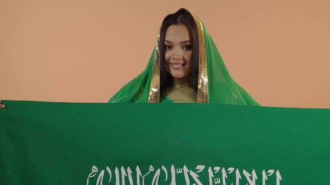 راية التوحيد وسيف القوة، السعادة بالانتماء للوطن، اليوم الوطني السعودي 23 سبتمبر، يوم العلم 11 مارس، ارتداء الزي الشعبي للمنطقة الوسطى في مناسبة وطنية, يوم بدينا22 فبراير, بورتريه مقرب لامرأة عربية خليجية سعودية ترتدي الشيلة والبشت تحمل علم المملكة، خلفية زهرية