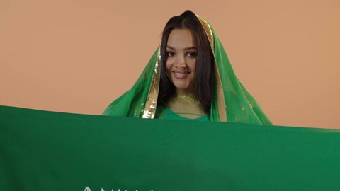 السعادة بالانتماء للوطن، راية التوحيد وسيف القوة، اليوم الوطني السعودي 23 سبتمبر، يوم العلم 11 مارس، ارتداء الزي الشعبي للمنطقة الوسطى في مناسبة وطنية, يوم بدينا22 فبراير, بورتريه مقرب لامرأة عربية خليجية سعودية ترتدي الشيلة والبشت تحمل علم المملكة، خلفية زهرية