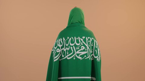 The concept of patriotism and belonging to the homeland, celebrating Saudi National Day on September 23, Flag Day on March 11, wearing the traditional costume of the central region on a national occasion, a portrait from the back of a Saudi Arabian Gulf woman wearing a green shayla and bisht, placing the Kingdom’s flag on her shoulders, a floral background