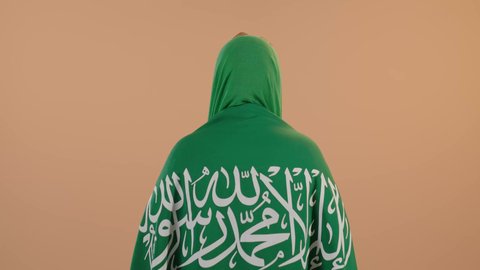Love of the homeland and belonging to it, celebrating Saudi National Day on September 23, Flag Day on March 11, wearing the traditional costume of the central region on a national occasion, a portrait from the back of a Saudi Arabian Gulf woman wearing a green shayla and bisht, placing the Kingdom’s flag on her shoulders, a floral background