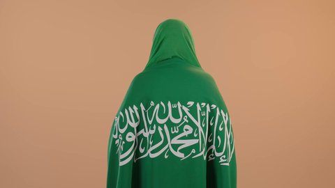 The concept of patriotism and belonging to the homeland, celebrating Saudi National Day on September 23, Flag Day on March 11, wearing the traditional costume of the central region on a national occasion, a portrait from the back of a Saudi Arabian Gulf woman wearing a green shayla and bisht, placing the Kingdom’s flag on her shoulders, a floral background