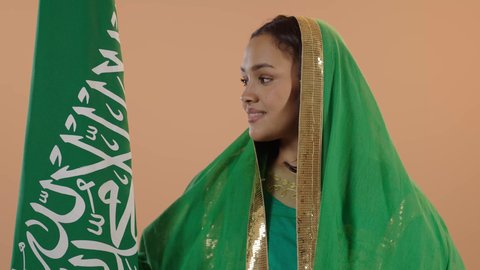 راية التوحيد وسيف القوة، السعادة بالانتماء للوطن، اليوم الوطني السعودي 23 سبتمبر، يوم العلم 11 مارس، ارتداء الزي الشعبي للمنطقة الوسطى في مناسبة وطنية, يوم بدينا22 فبراير, بورتريه مقرب لامرأة عربية خليجية سعودية ترتدي الشيلة والبشت تحمل علم المملكة، خلفية زهرية