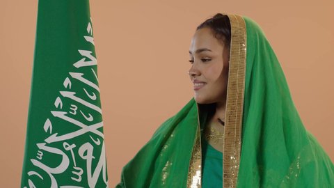 السعادة بالانتماء للوطن، راية التوحيد وسيف القوة، اليوم الوطني السعودي 23 سبتمبر، يوم العلم 11 مارس، ارتداء الزي الشعبي للمنطقة الوسطى في مناسبة وطنية, يوم بدينا22 فبراير, بورتريه مقرب لامرأة عربية خليجية سعودية ترتدي الشيلة والبشت تحمل علم المملكة، خلفية زهرية