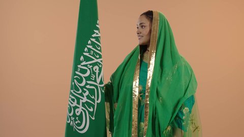 Boasting of the nation's achievements, the banner of monotheism and the sword of strength, Saudi National Day September 23, Flag Day March 11, wearing the traditional costume of the central region on a national occasion, Badina Day February 22, portrait of a Saudi Arabian Gulf woman wearing a sheila and bisht holding the flag of the Kingdom, floral background